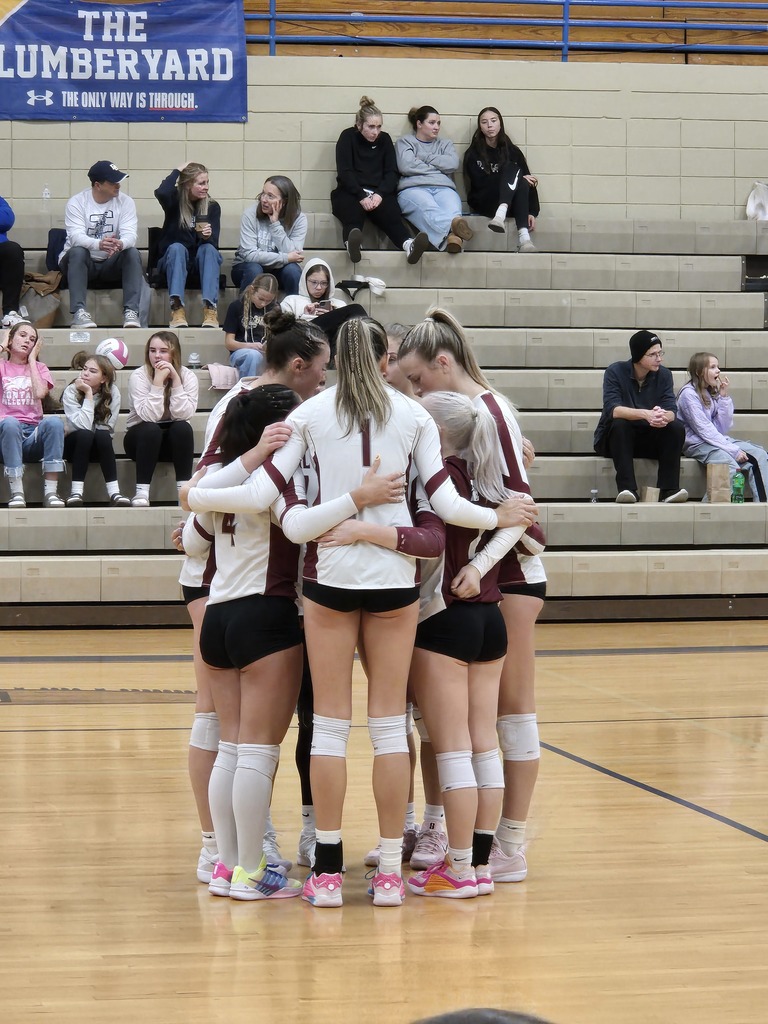 hamilton high school volleyball stands in a tight circle with their arms around each other on a volleyball court during a divisional game