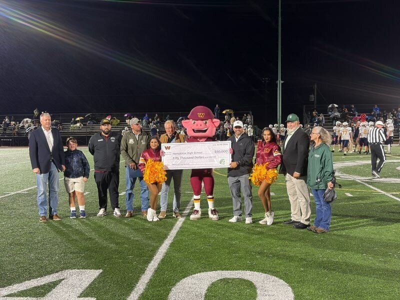 A group of school and community representatives, students, cheerleaders, and a mascot gather on the Hampshire High School football field for a $50,000 check presentation. The group holds a large ceremonial check while posing for a photograph during a nighttime football game.