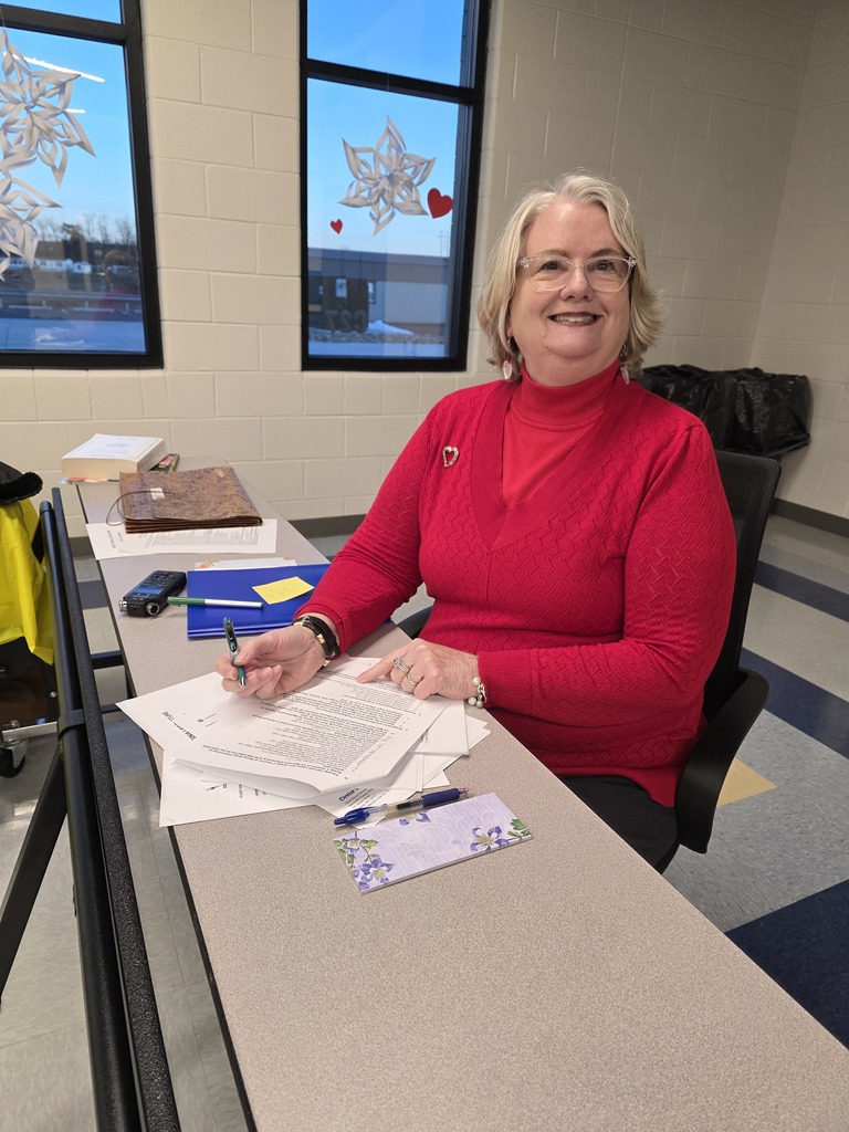 A smiling woman with short blonde hair and glasses sits at a table in a school setting, wearing a bright red sweater and a heart-shaped brooch. She is holding a pen and reviewing paperwork with a welcoming expression. The background features large windows with snowflake and heart decorations, adding a warm and festive touch to the room.
