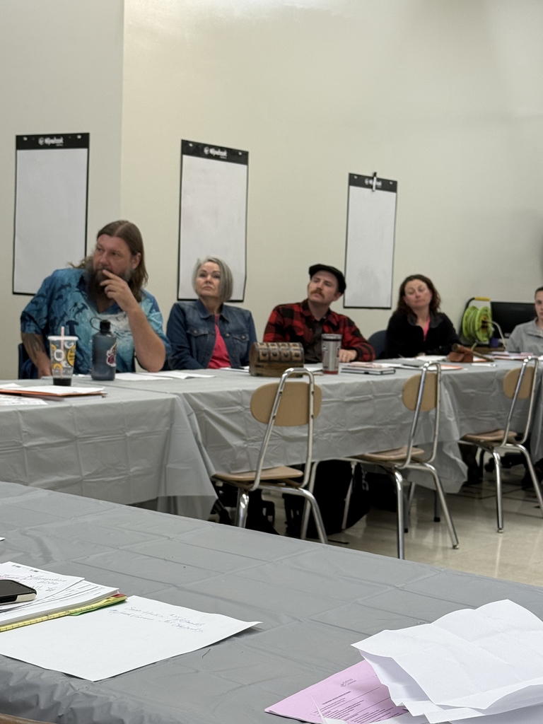 A group of adults seated around a large U-shaped arrangement of tables in a classroom setting, attending what appears to be a school-related training or professional development session. Tables are covered in gray plastic tablecloths and scattered with folders, handouts, notebooks, and drinks. Some participants are taking notes or listening attentively, while others are seated at computers along the wall. Posters, dry erase boards, and classroom fixtures are visible in the background, indicating a school environment. The atmosphere appears focused and collaborative.  