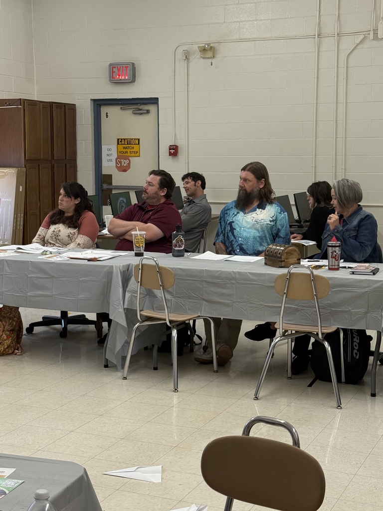 A group of adults seated around a large U-shaped arrangement of tables in a classroom setting, attending what appears to be a school-related training or professional development session. Tables are covered in gray plastic tablecloths and scattered with folders, handouts, notebooks, and drinks. Some participants are taking notes or listening attentively, while others are seated at computers along the wall. Posters, dry erase boards, and classroom fixtures are visible in the background, indicating a school environment. The atmosphere appears focused and collaborative.  
