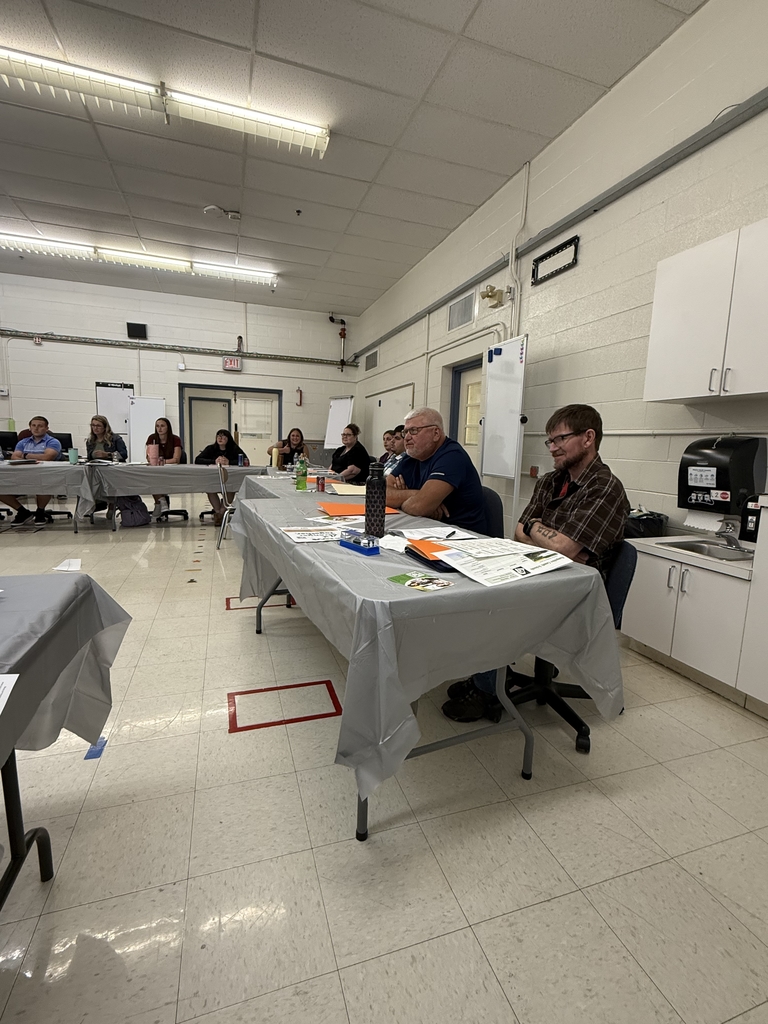 A group of adults seated around a large U-shaped arrangement of tables in a classroom setting, attending what appears to be a school-related training or professional development session. Tables are covered in gray plastic tablecloths and scattered with folders, handouts, notebooks, and drinks. Some participants are taking notes or listening attentively, while others are seated at computers along the wall. Posters, dry erase boards, and classroom fixtures are visible in the background, indicating a school environment. The atmosphere appears focused and collaborative.  