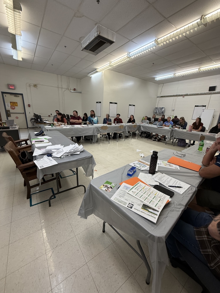 A group of adults seated around a large U-shaped arrangement of tables in a classroom setting, attending what appears to be a school-related training or professional development session. Tables are covered in gray plastic tablecloths and scattered with folders, handouts, notebooks, and drinks. Some participants are taking notes or listening attentively, while others are seated at computers along the wall. Posters, dry erase boards, and classroom fixtures are visible in the background, indicating a school environment. The atmosphere appears focused and collaborative.

