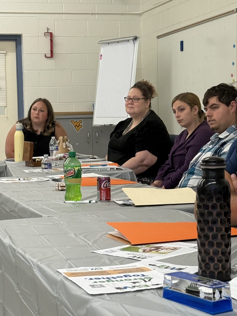 A group of adults seated around a large U-shaped arrangement of tables in a classroom setting, attending what appears to be a school-related training or professional development session. Tables are covered in gray plastic tablecloths and scattered with folders, handouts, notebooks, and drinks. Some participants are taking notes or listening attentively, while others are seated at computers along the wall. Posters, dry erase boards, and classroom fixtures are visible in the background, indicating a school environment. The atmosphere appears focused and collaborative.  