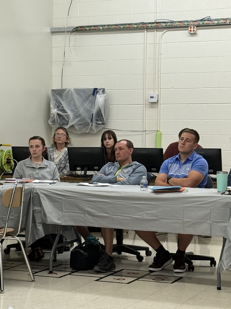 A group of adults seated around a large U-shaped arrangement of tables in a classroom setting, attending what appears to be a school-related training or professional development session. Tables are covered in gray plastic tablecloths and scattered with folders, handouts, notebooks, and drinks. Some participants are taking notes or listening attentively, while others are seated at computers along the wall. Posters, dry erase boards, and classroom fixtures are visible in the background, indicating a school environment. The atmosphere appears focused and collaborative.  