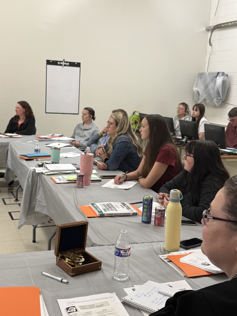 A group of adults seated around a large U-shaped arrangement of tables in a classroom setting, attending what appears to be a school-related training or professional development session. Tables are covered in gray plastic tablecloths and scattered with folders, handouts, notebooks, and drinks. Some participants are taking notes or listening attentively, while others are seated at computers along the wall. Posters, dry erase boards, and classroom fixtures are visible in the background, indicating a school environment. The atmosphere appears focused and collaborative.  
