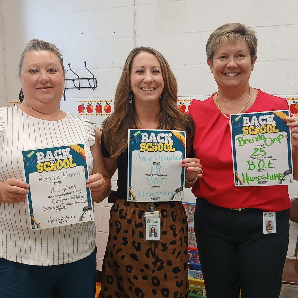 Three women standing together and smiling, each holding “Back to School” certificates. The woman on the left is wearing a striped blouse, the middle woman is wearing a black top with a leopard-print skirt, and the woman on the right is wearing a red blouse.