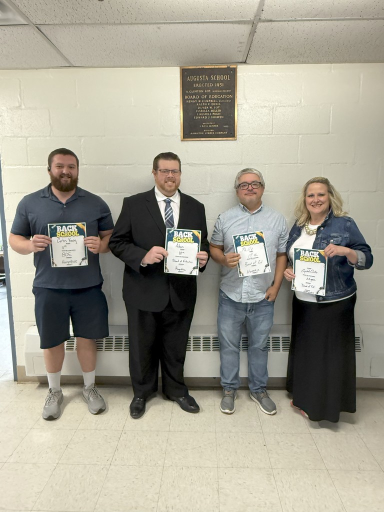 Four people standing side by side in front of a wall plaque at Augusta School, each holding “Back to School” certificates. From left to right, a man in a polo shirt and shorts, a man in a suit, a man in a button-up shirt, and a woman in a long skirt and denim jacket.