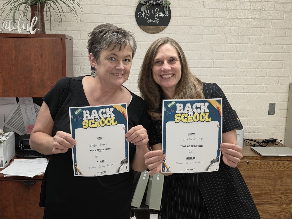 Two smiling Board of Education employees holding “Back to School” certificates. The woman on the left is wearing a black top with earrings, and the woman on the right is wearing a black striped dress.