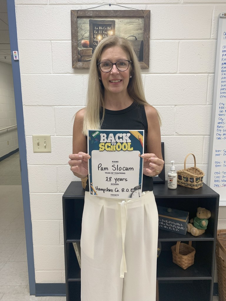 A woman standing indoors, smiling and holding a “Back to School” certificate. She is wearing glasses, a sleeveless black blouse, and white pants.
