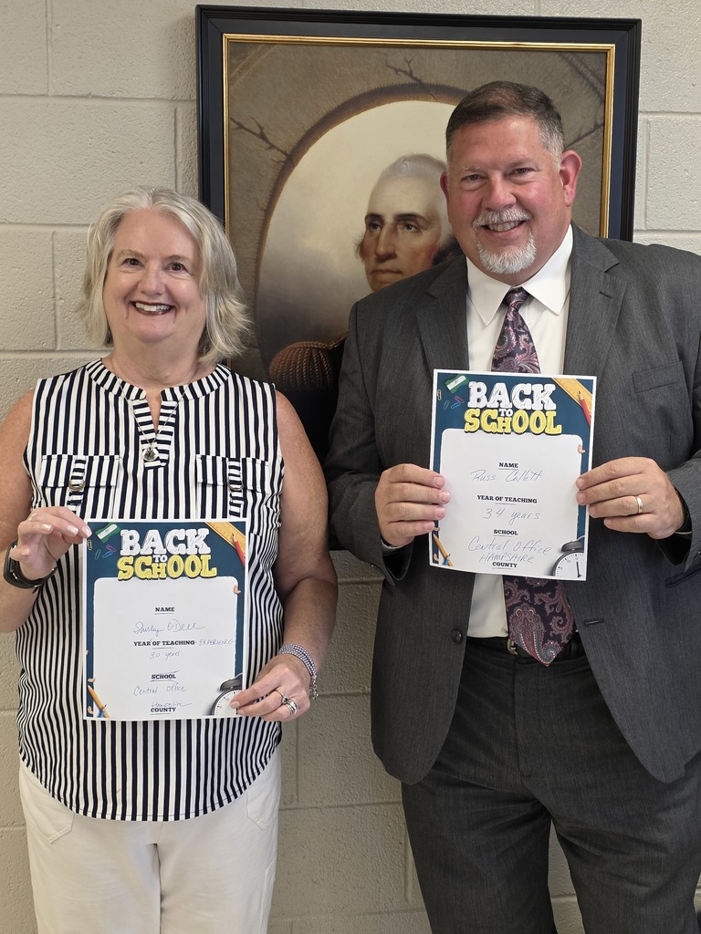 Two people smiling and holding “Back to School” certificates in front of a framed portrait of George Washington. The woman on the left is wearing a striped blouse, and the man on the right is wearing a suit and tie.