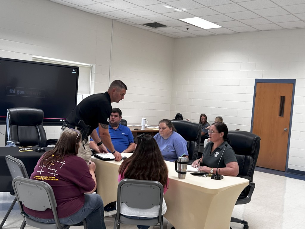 A man in a dark shirt speaks while gesturing with his hands at a table surrounded by staff members. Others sit with focused expressions, listening intently to the scenario being discussed.