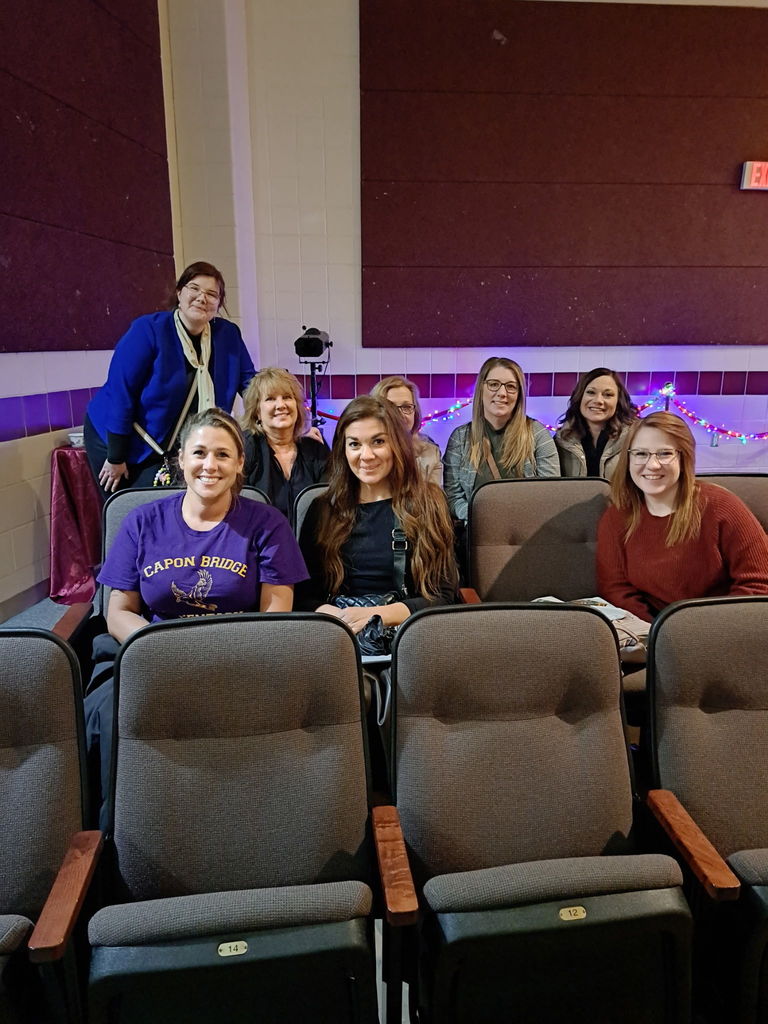 A group of seven Hampshire County Schools staff members are seated together in an auditorium, smiling for a photo during the Communities In Schools “Keep the Ball Rolling” event. The women in the front row wear casual attire, including a purple Capon Bridge shirt, while those in the back row sit against a maroon and white wall decorated with string lights. The atmosphere is friendly and collaborative, highlighting the educators’ participation in a community partnership event focused on supporting student success.