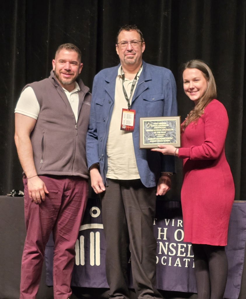 Aaron Henderson stands on stage holding a plaque recognizing him as Middle School Counselor of the Year. He is joined by two presenters. A West Virginia School Counselor Association banner and stage backdrop are visible behind them.