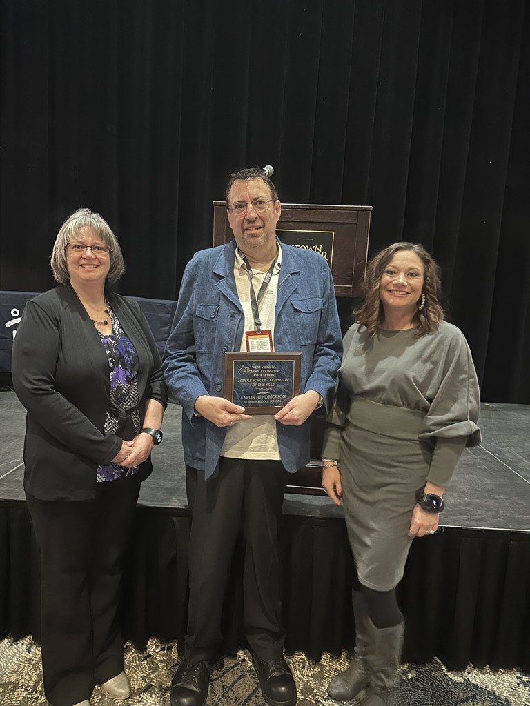 Aaron Henderson stands on stage holding his Middle School Counselor of the Year plaque, flanked by two colleagues. A podium and black stage curtain are visible behind them.