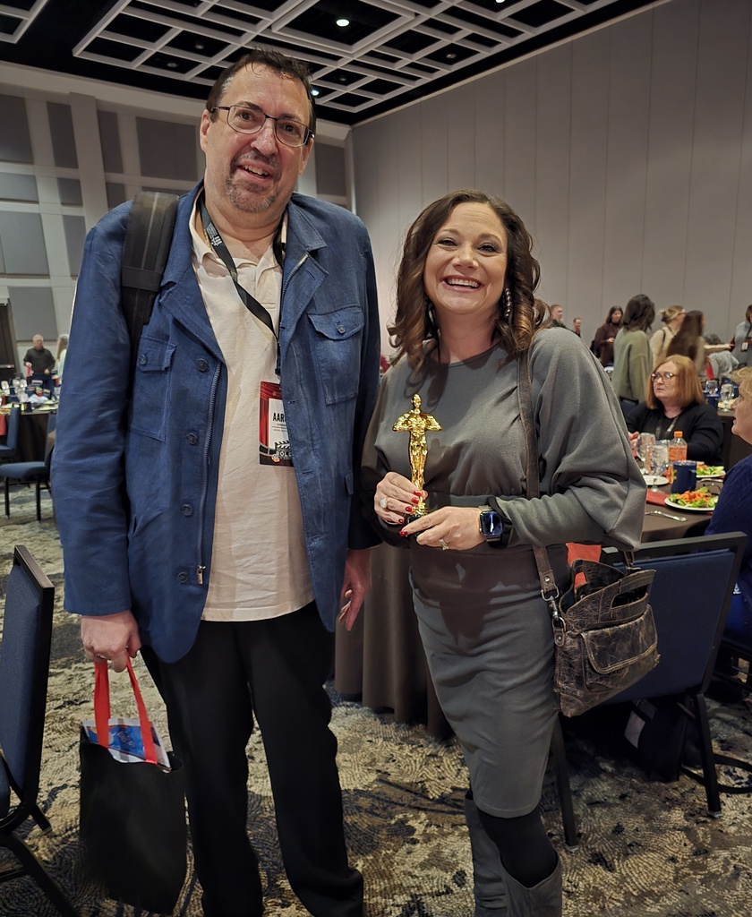 Aaron Henderson stands with a smiling colleague at an awards banquet. The colleague is holding a gold trophy. Round banquet tables and attendees are visible in the background.