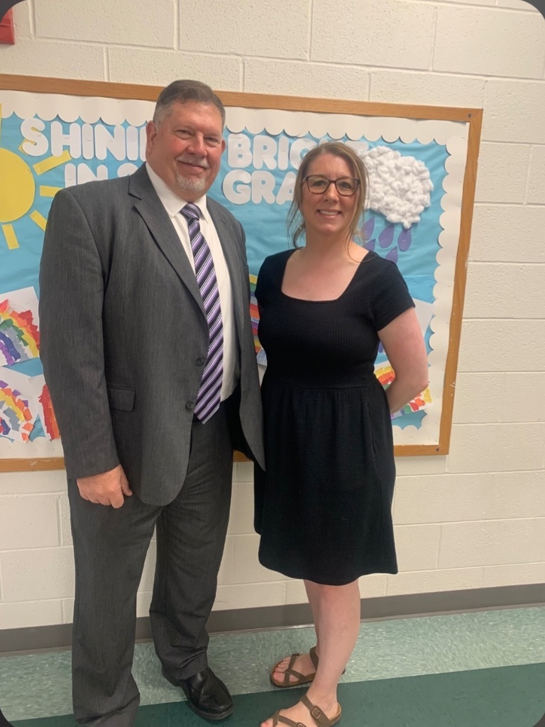 Two staff members stand in a school hallway in front of a bulletin board decorated with a sun, clouds, and student artwork. Both are smiling and dressed professionally.