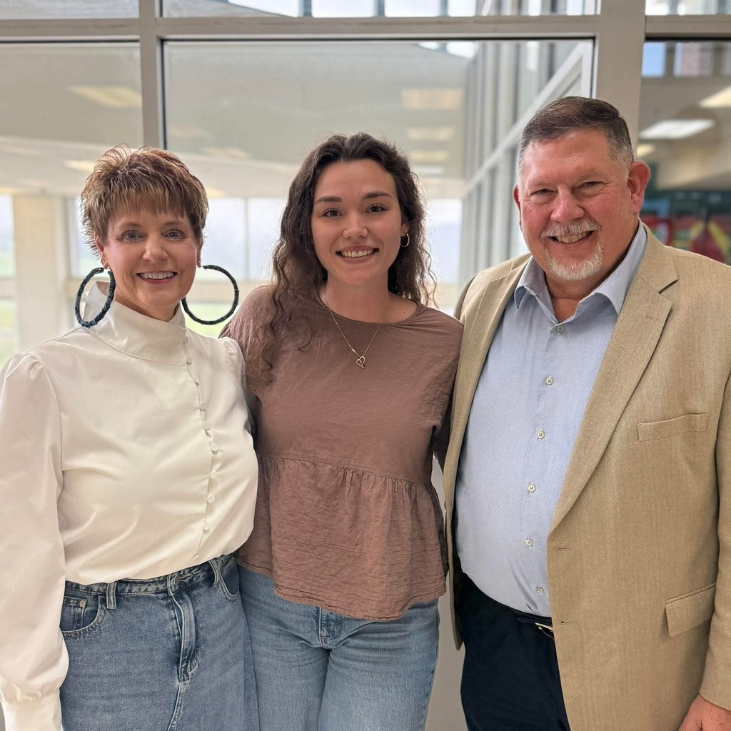Three staff members stand together indoors in front of large windows. They are smiling and standing shoulder to shoulder in a bright, open school space.