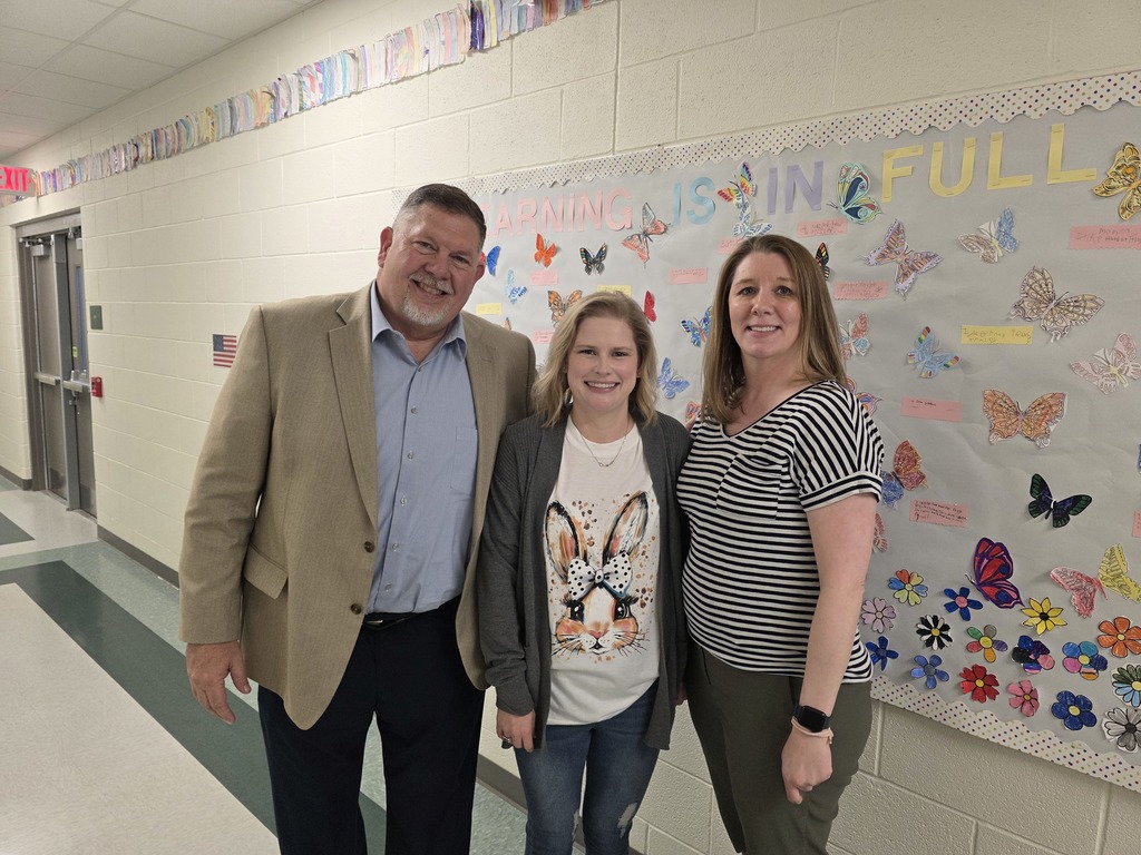 Three staff members stand in a school hallway in front of a bulletin board that reads “Learning is in Full Bloom,” decorated with colorful student-made butterflies. All three are smiling and standing close together.