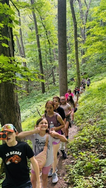 Students continue hiking single file along a forest trail. One student near the front smiles and gives a thumbs-up toward the camera while others follow behind among tall trees and dense greenery.