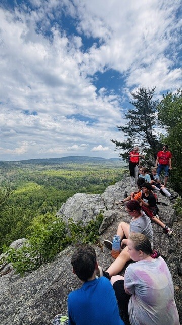 A group of students and adults sit and stand on a rocky overlook at the top of a mountain. They face outward, looking across a wide green forest valley under a partly cloudy blue sky.