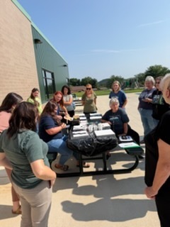A group of Hampshire County Schools staff members gather around picnic tables outside a school building during professional development. Staff are standing and seated together participating in an outdoor training activity.