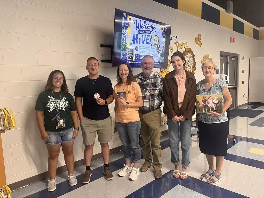 Six school staff members stand together and smile for a photo in a school hallway. Behind them, a display reads “Welcome Back to the Hive,” with bee-themed decorations welcoming staff back to school.