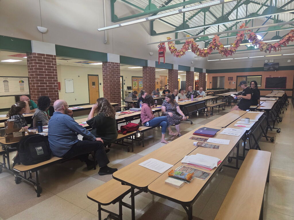 Staff members gather in small groups around cafeteria tables during a professional development session. Training materials are spread across the tables as participants talk and work together.