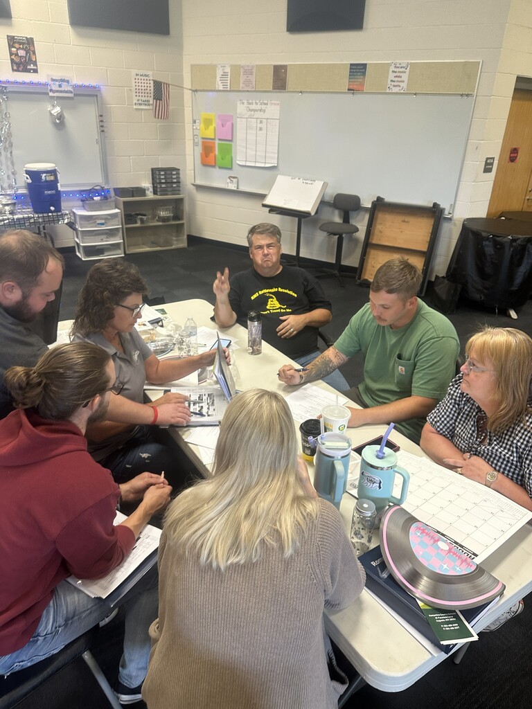 School staff members sit together around a classroom table covered with papers, notebooks, drinks, and training materials. The group is engaged in discussion and collaborative professional learning.