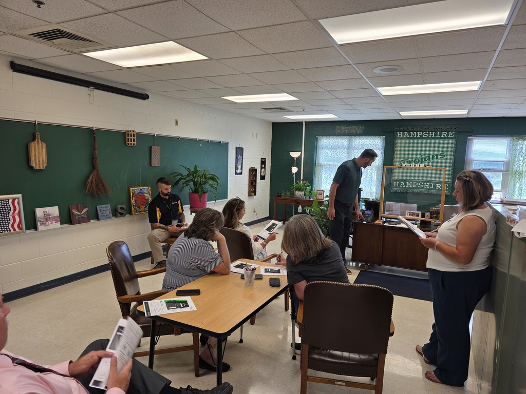 A small group of staff members participate in professional development inside a classroom decorated in school colors. Several participants are seated around a table while others stand and lead or participate in the discussion.