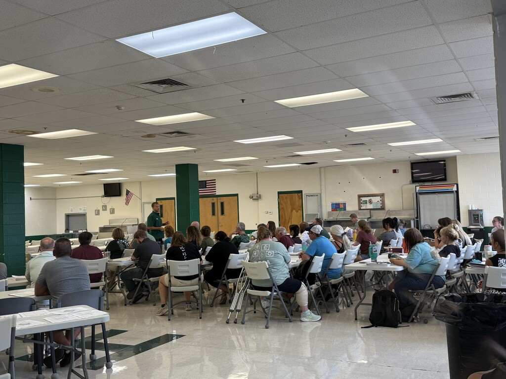 A large group of Hampshire County Schools employees participate in professional development in a school cafeteria. Staff members sit at tables facing a presenter at the front of the room while reviewing training materials.