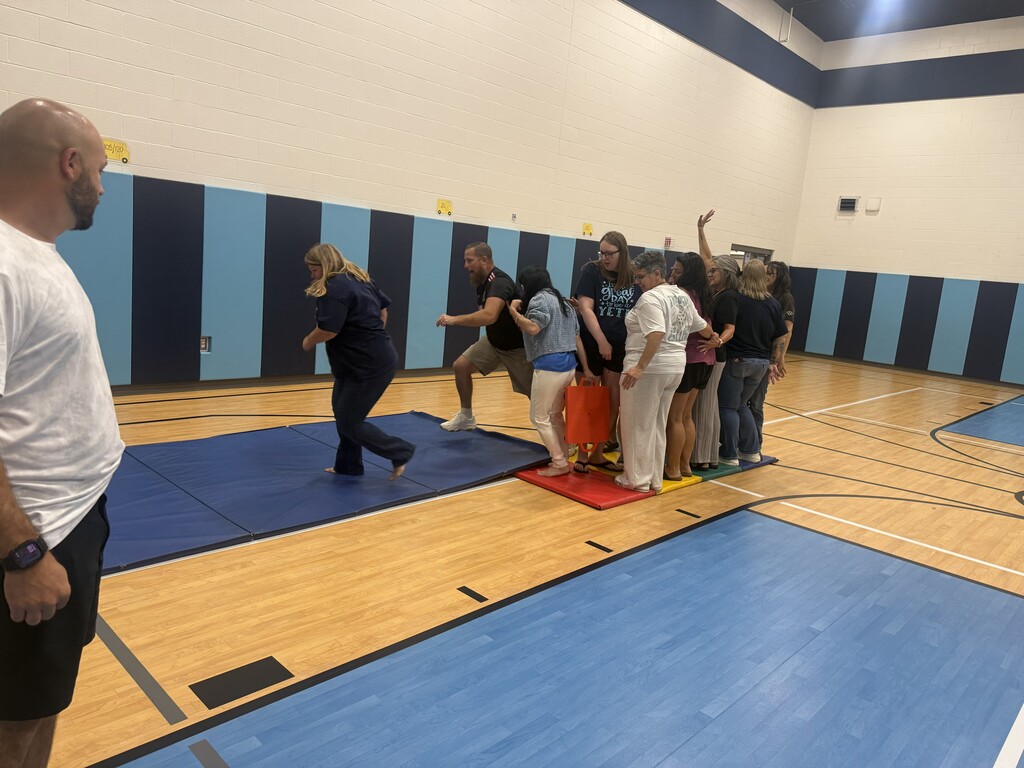 School staff members participate in an interactive team-building activity in a gymnasium. The group works closely together on colorful mats while other staff members assist and observe.