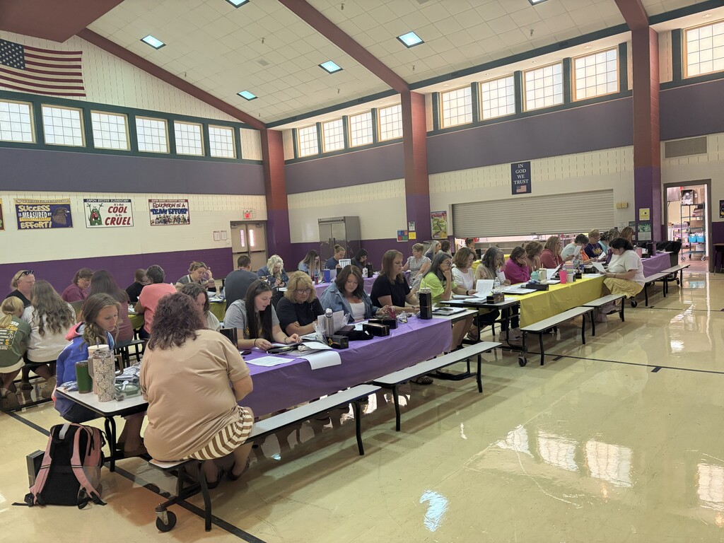 A large group of school staff members sit together at long tables in an elementary school gymnasium. Participants are reading materials and working together during a professional development session.