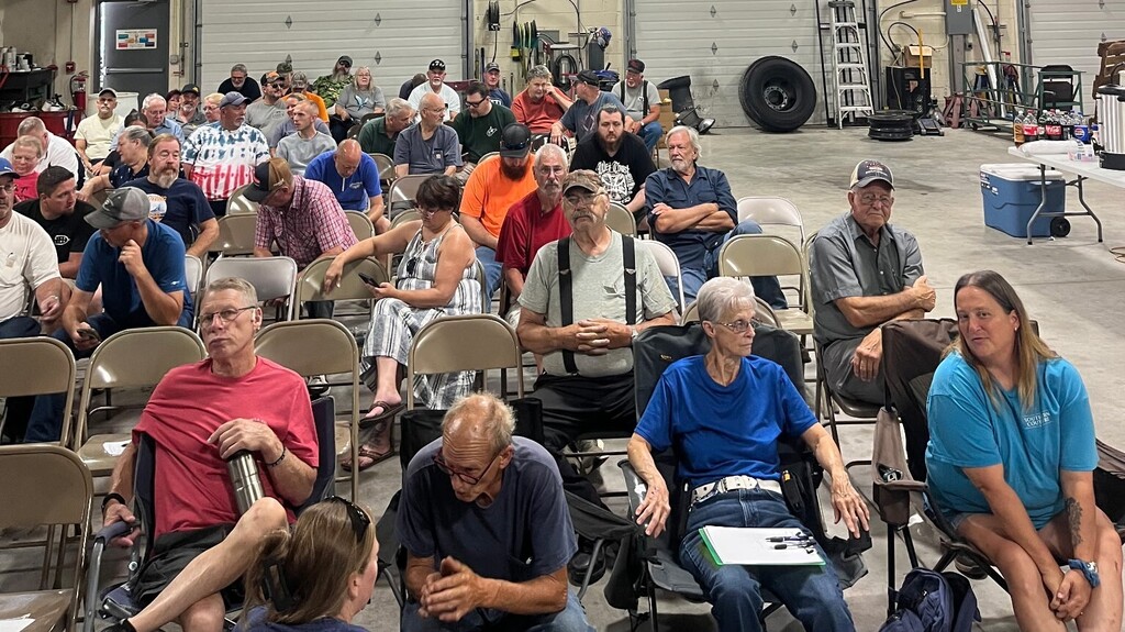 A large group of Hampshire County Schools bus drivers and transportation staff seated in rows of chairs inside a transportation garage during a back-to-school training session. Staff members are gathered together listening, talking, and preparing for the upcoming school year. School transportation equipment and garage doors are visible in the background.