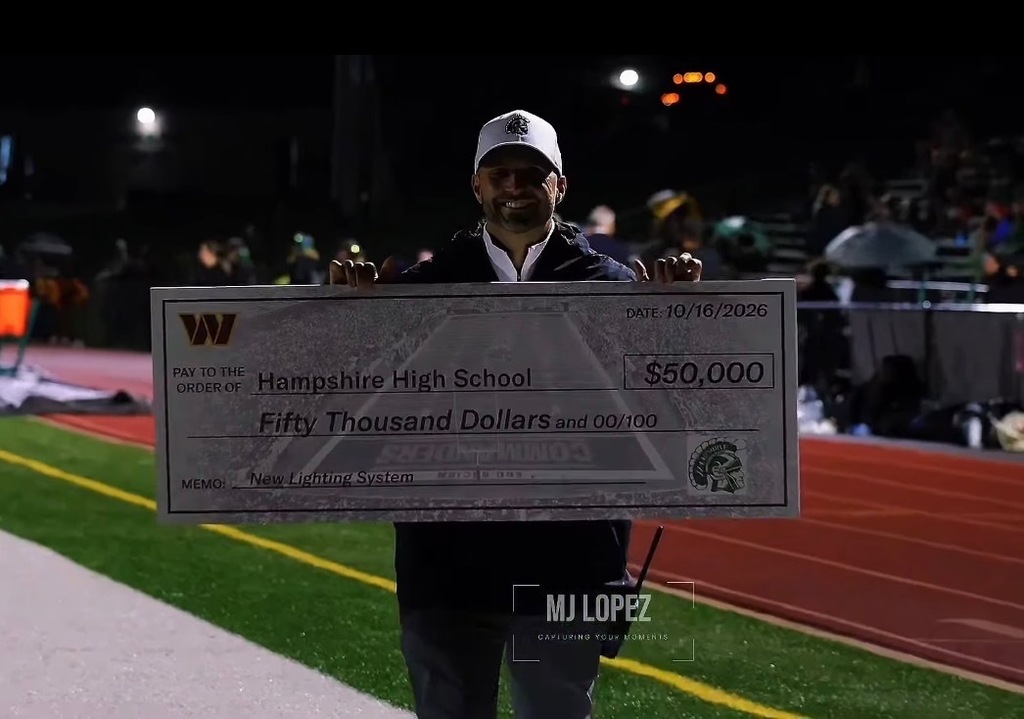 A man wearing a Hampshire Trojans hat holds a large ceremonial check for $50,000 made payable to Hampshire High School. The check, dated October 16, 2026, identifies the donation as funding for a new lighting system. The photograph was taken on the football field during an evening event.
