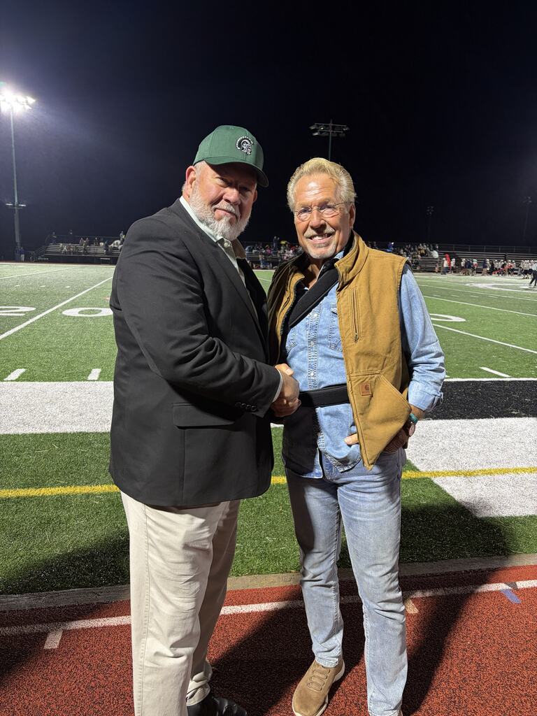 Two men shake hands on the Hampshire High School football field during an evening event. One wears a black blazer, khaki pants, and a green Hampshire Trojans hat. The other wears a tan vest over a blue denim shirt. Stadium lights illuminate the field in the background.