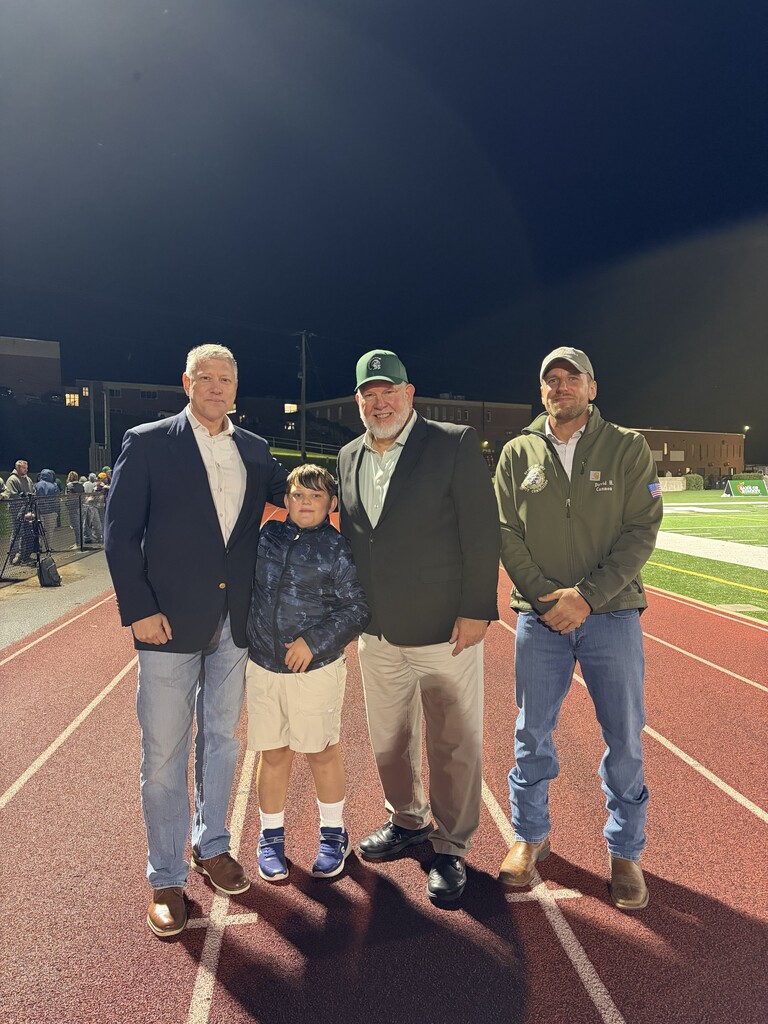 Four people, including three men and a young boy, pose together on the Hampshire High School track during a nighttime football game. The group stands in front of the football field, with stadium lights and school buildings visible in the background.