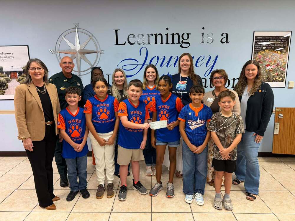a group of students wearing matching odyssey of the mind t-shirts, with several adults, holding a donation check