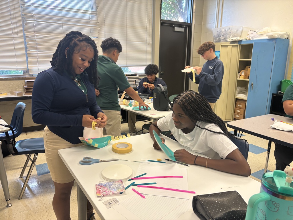 Students and an adult in a classroom, working on crafts. Two women seated at a table, with various craft supplies and scissors.