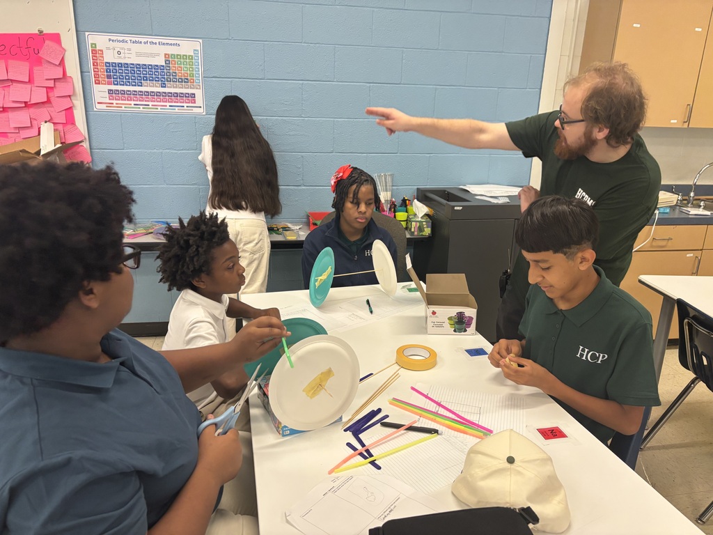 Several children work on crafts at a table in a classroom. A man in a green shirt stands behind them.