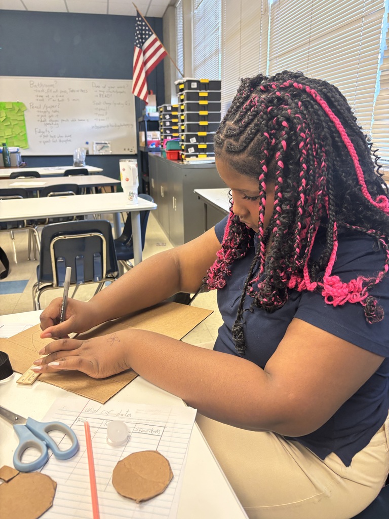 A person works on paper crafts at a table in a classroom. A flag and a whiteboard are in the background.