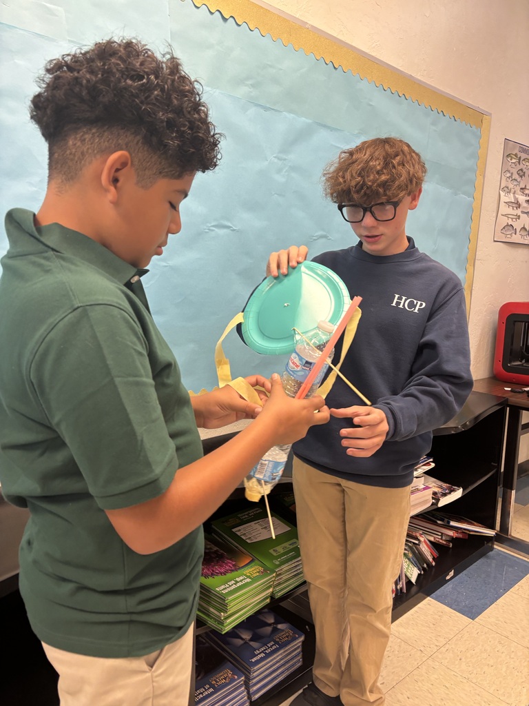 Two boys in a classroom, one holding a green object, both examining it closely. Behind them, a bulletin board and shelves with books.