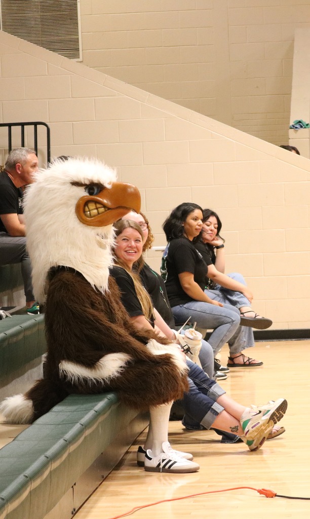 A person in a costume sits on a bench. Other people sit behind them in a gym.