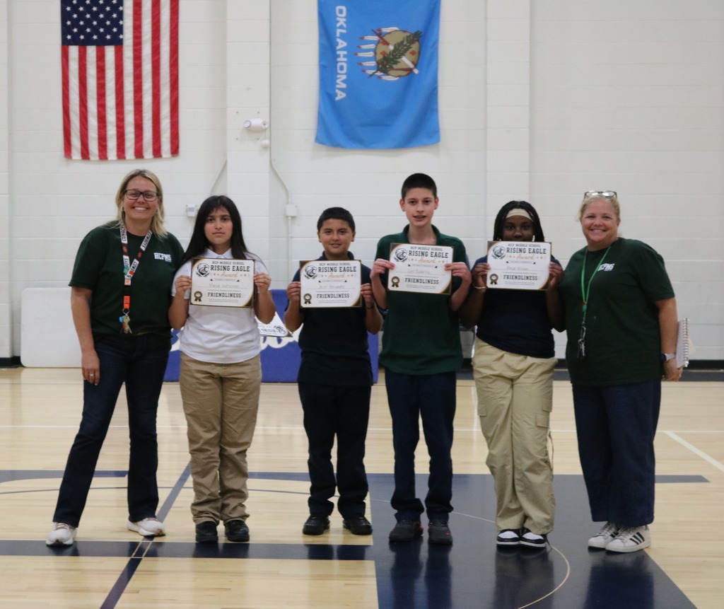 Six people stand on a basketball court, holding certificates. They wear matching green shirts and pants. Two flags hang in the background.