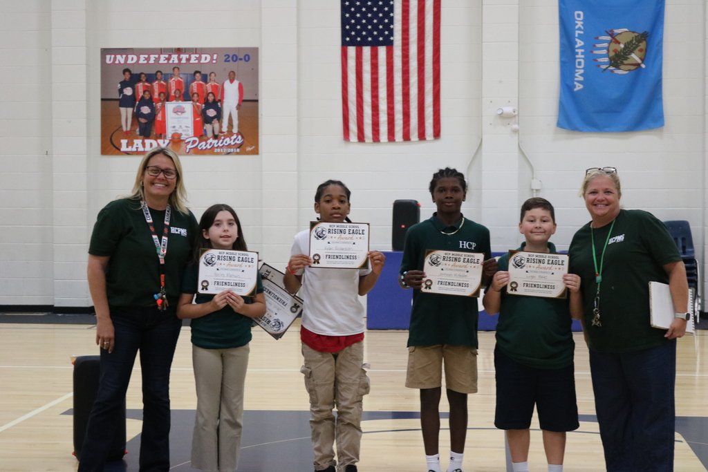 A group of children and adults stand in a gym. They hold certificates. The background includes a blue flag and an American flag.