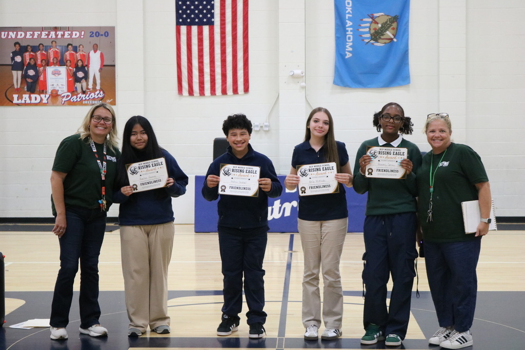 Five individuals stand on a basketball court, holding certificates, with two flags hanging on the wall.