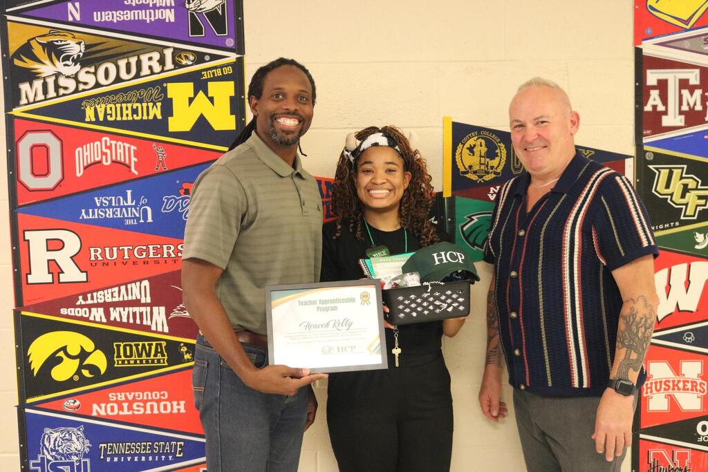 Three individuals stand before a wall displaying banners from various universities. A woman holds a certificate and a purse.