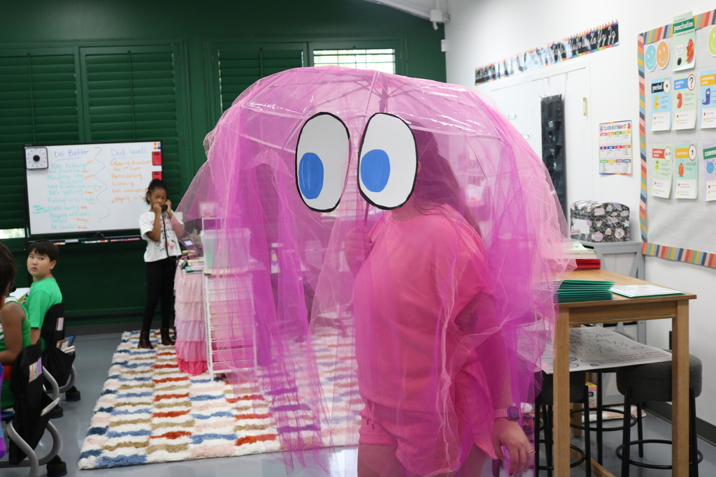 A person in a pink outfit with a large pink umbrella with large eyes stands in a classroom.