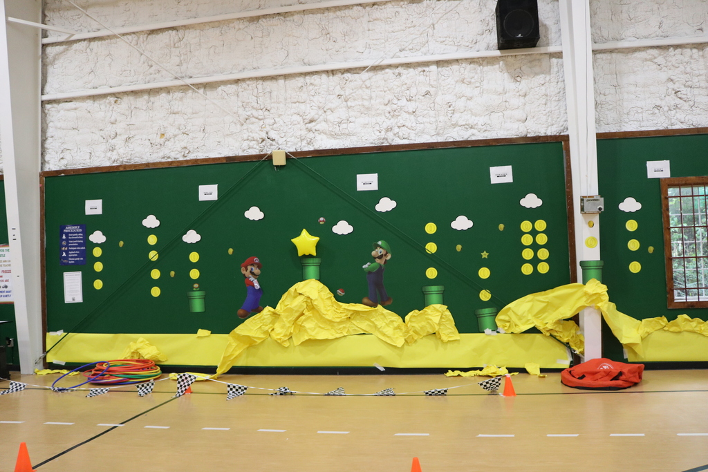 Green backdrop with yellow and white elements, characters, and a yellow platform. Cones line the floor.