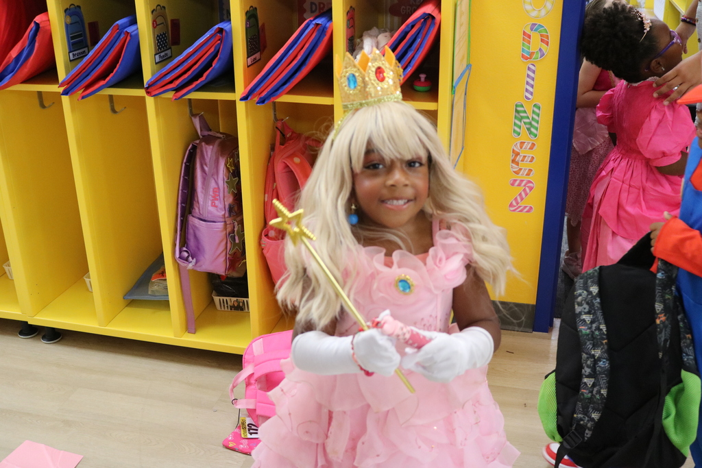A young girl in a pink dress and blonde wig, wearing a crown and holding a wand, stands near yellow cubbies.
